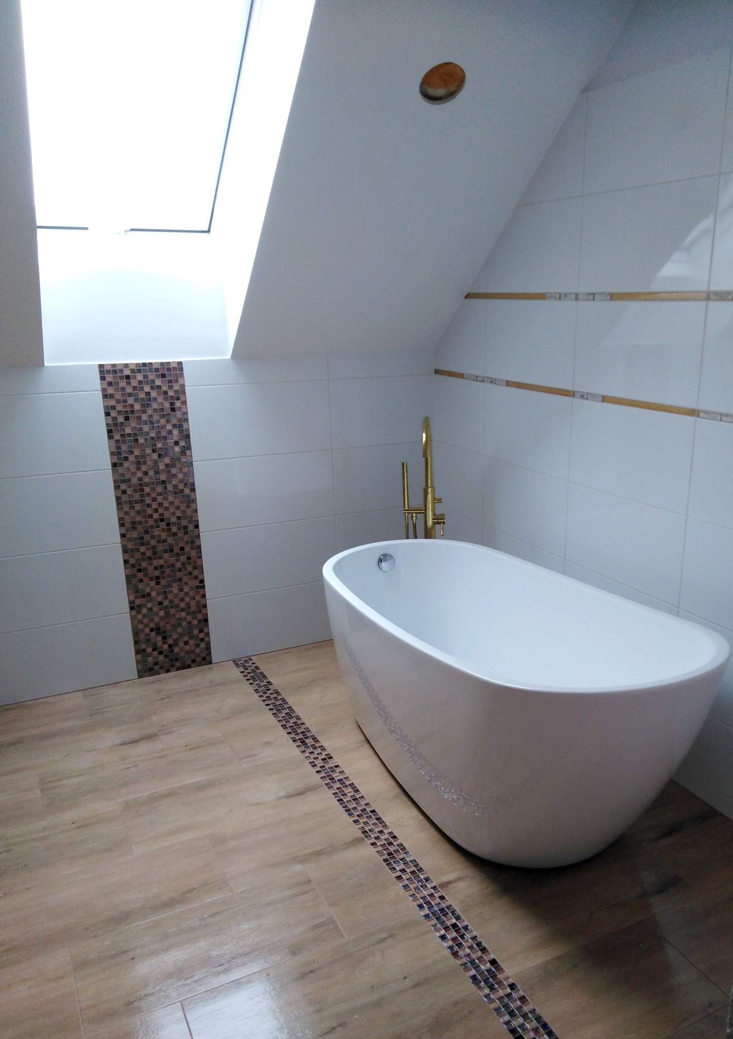 Nowoczesna łazienka na poddaszu z wolnostojącą białą wanną, złotą armaturą, wykończona białymi płytkami z dekoracyjnymi paskami mozaiki i oknem dachowym.