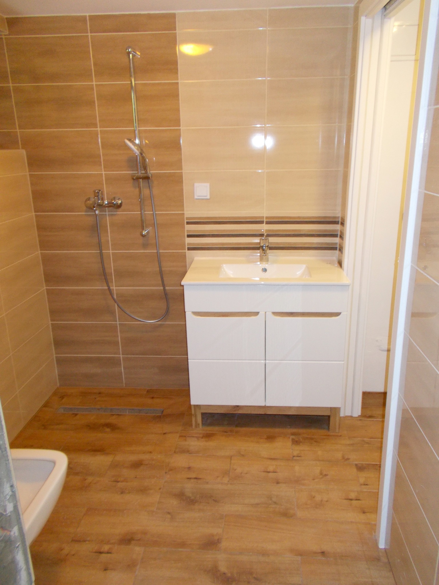 Wykończona łazienka z brązowymi płytkami imitującymi drewno na podłodze i ścianach, biała szafka pod umywalką z dwiema szafkami, chromowana bateria i deszczownica, widoczny fragment toalety.