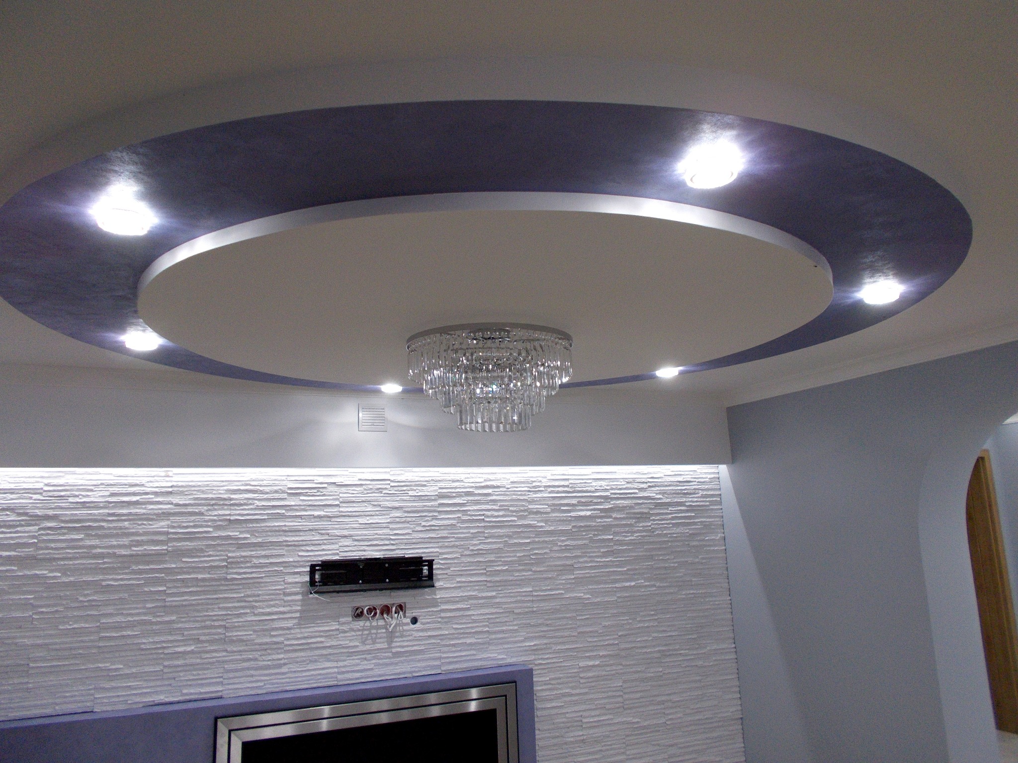 Sufit podwieszany z okrągłym, białym elementem centralnym i szarą obwódką, oświetlony punktowymi lampami i kryształowym żyrandolem, ściana pokryta dekoracyjną, białą strukturą przypominającą kamień.