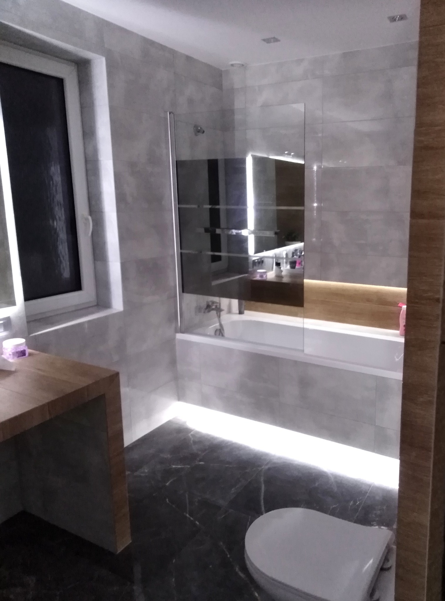 Nowoczesna łazienka z szarymi płytkami imitującymi kamień, podświetlaną wanną z parawanem, blatem z drewna i czarną marmurową podłogą.