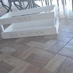 Biała, dwupoziomowa skrzynka z napisem 'WINO' i 'DLA MŁODYCH' stoi na podłodze z płytek imitujących drewno, w tle widać metalowe nogi stołu i białą zasłonę.