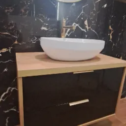 Nowoczesna łazienka z umywalką nablatową w kształcie misy, złotą armaturą i okrągłym lustrem na tle ściany z czarnego marmuru.