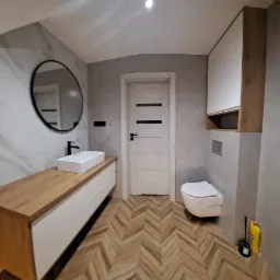 Nowoczesna łazienka z drewnianym blatem pod umywalką, owalnym lustrem w czarnej ramie, białymi szafkami wiszącymi i podwieszaną toaletą, wykończona płytkami imitującymi marmur i drewnianą podłogą...