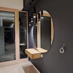 Nowoczesna toaletka z okrągłym lustrem i drewnianym blatem zamontowana na ciemnoszarej ścianie, oświetlona wiszącą lampą z sześcioma czarnymi reflektorami, widok na przeszklone drzwi.