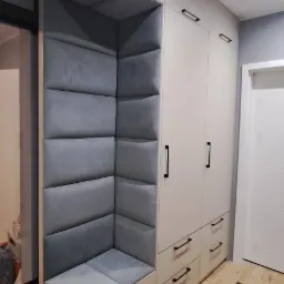 Biała szafa wnękowa z siedziskiem i tapicerowanym oparciem w kolorze szarym, czarne uchwyty, jasna podłoga.