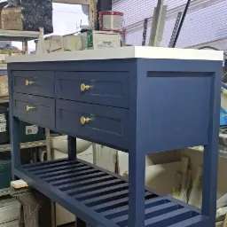 Niebieska szafka łazienkowa z czterema szufladami i złotymi uchwytami, stojąca na kozłach w warsztacie, z widocznym regałem pod blatem i białym blatem.