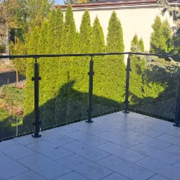 Szklana balustrada balkonowa z czarnymi, metalowymi słupkami o przekroju krzyża, widok na zieleń i budynki w tle.