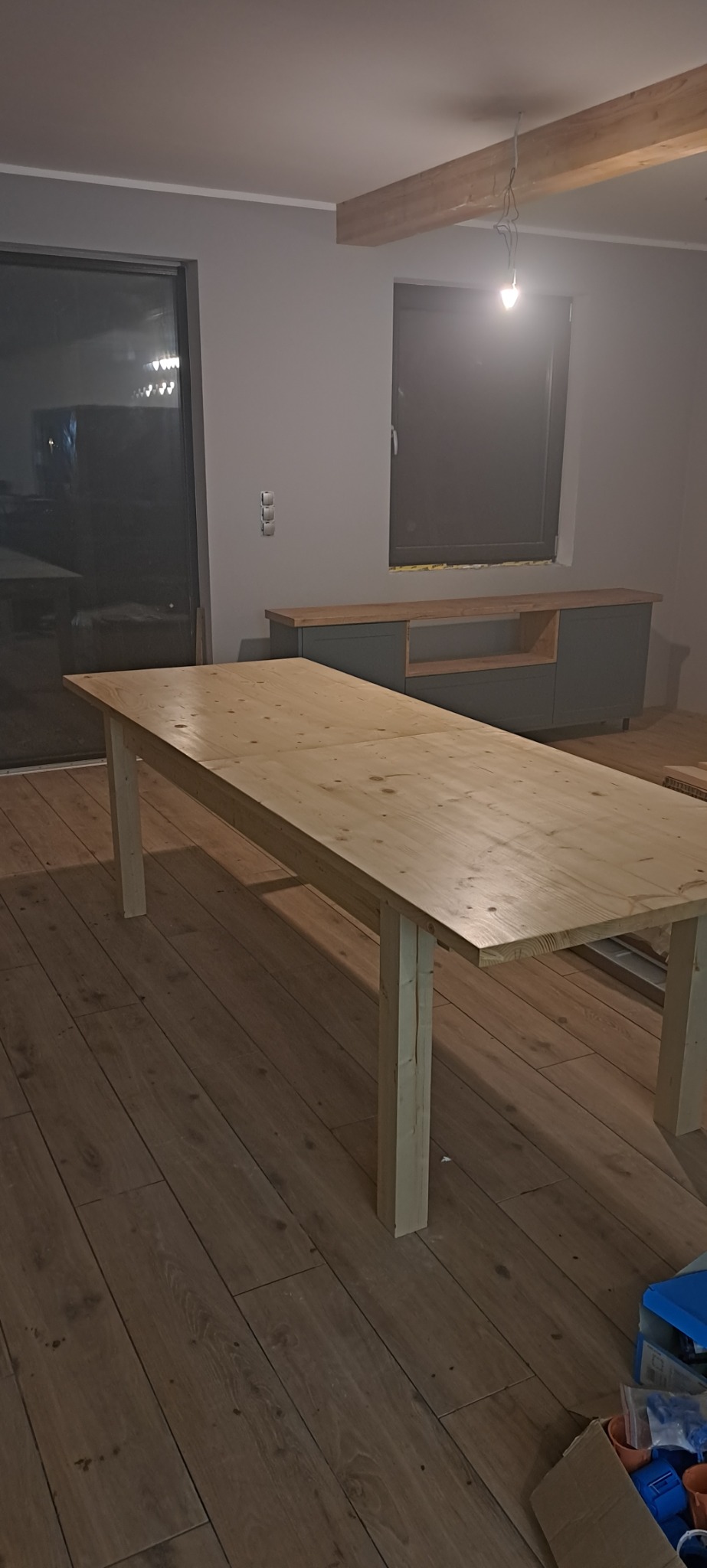 Surowy, jasny stół z drewna sosnowego na tle szarej ściany z oknem i komodą, widoczne niedokończone wykończenie wnętrza z drewnianą belką stropową i wystającymi kablami.