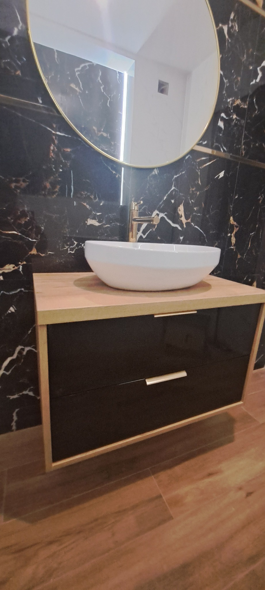 Nowoczesna łazienka z umywalką nablatową w kształcie misy, złotą armaturą i okrągłym lustrem na tle ściany z czarnego marmuru.
