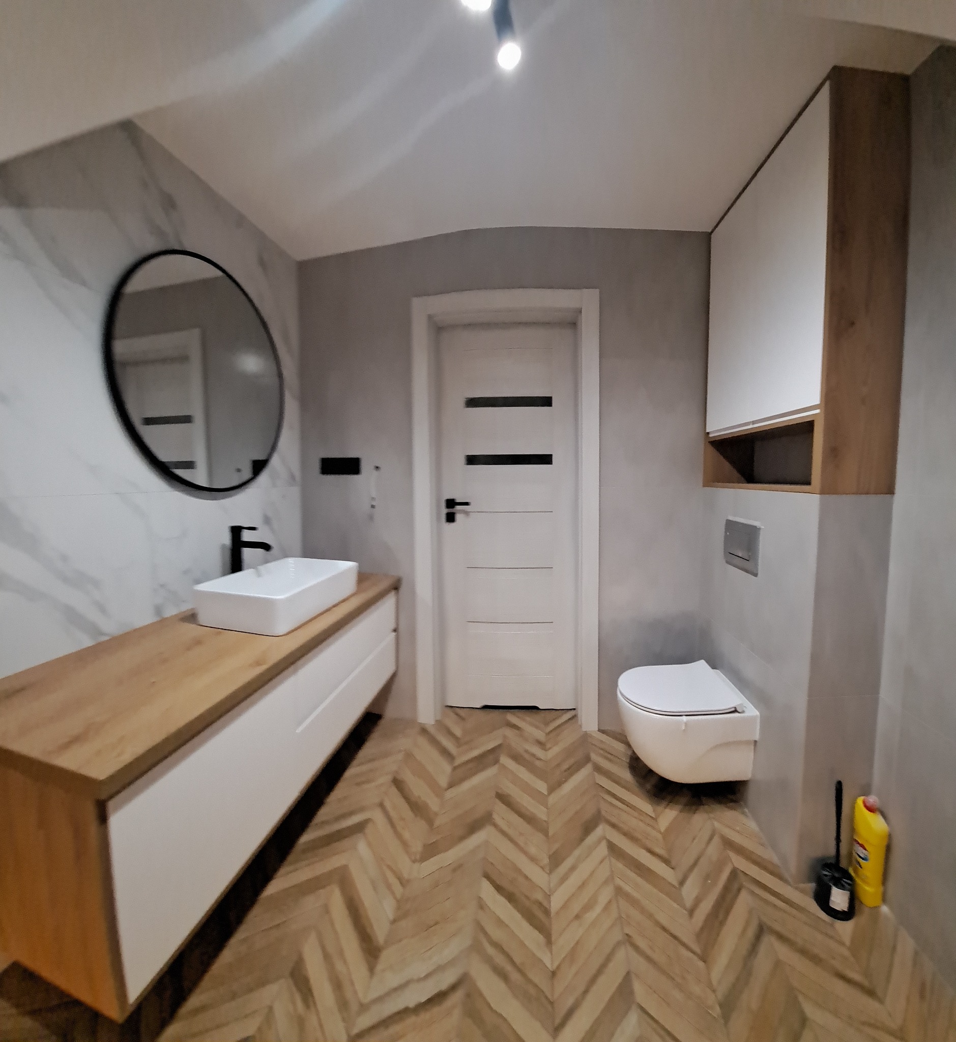 Nowoczesna łazienka z drewnianym blatem pod umywalką, owalnym lustrem w czarnej ramie, białymi szafkami wiszącymi i podwieszaną toaletą, wykończona płytkami imitującymi marmur i drewnianą podłogą...