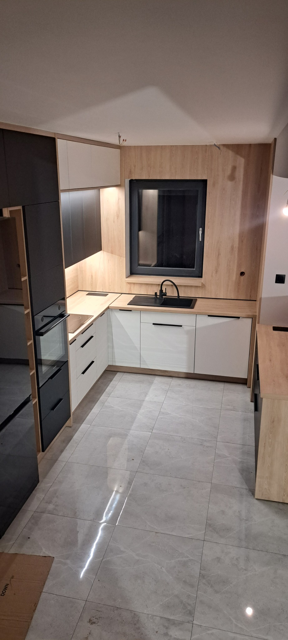 Nowoczesna kuchnia w jasnych barwach z białymi szafkami, drewnianymi blatami i czarnymi akcentami, w tym zlewem i oknem. Widoczne niedokończone wykończenie ściany z wystającymi kablami.