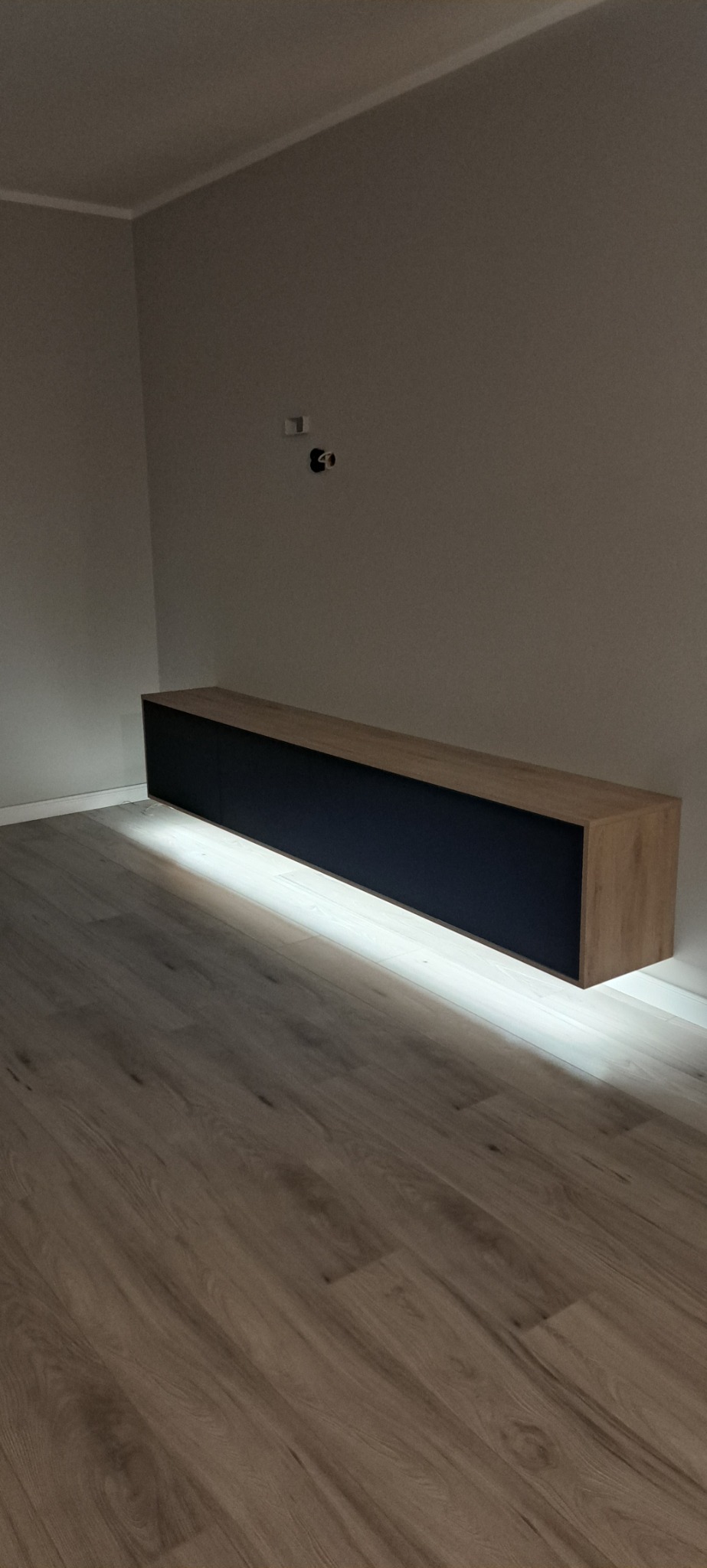 Długa, wisząca szafka RTV w kolorze dębu i granatu, podświetlona od dołu listwą LED, zawieszona na szarej ścianie nad podłogą z paneli.