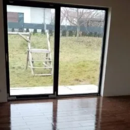 Pokój w stanie surowym z nową, błyszczącą podłogą z paneli, widoczne ślady po pracach wykończeniowych na ścianach, duże okno przesuwne z widokiem na ogród i drewnianą konstrukcję drabiny.