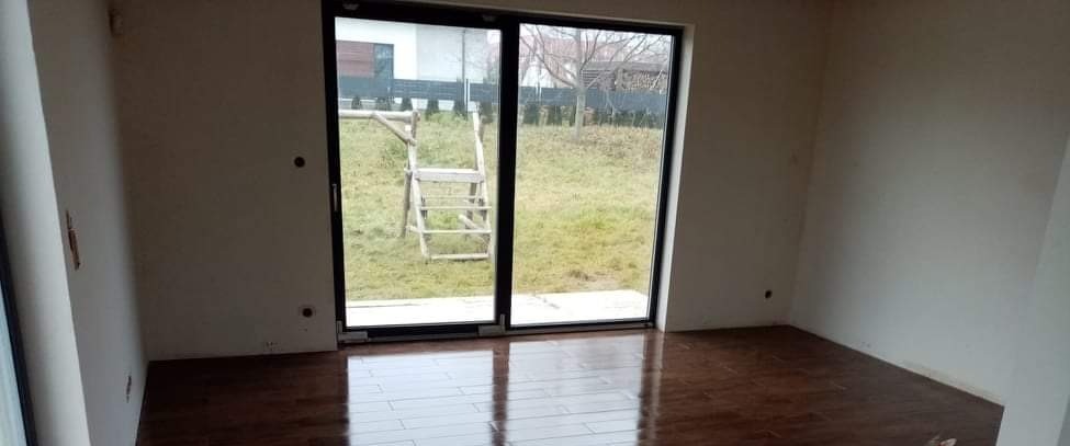 Pokój w stanie surowym z nową, błyszczącą podłogą z paneli, widoczne ślady po pracach wykończeniowych na ścianach, duże okno przesuwne z widokiem na ogród i drewnianą konstrukcję drabiny.