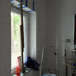 ROMIX Wykończenia wnętrza - Ściana łazienki w trakcie wykańczania, z białymi płytkami, oknem zabezpieczonym taśmą malarską, opartą deską, butelką napoju, ubraniem na parapecie i aluminiową drabiną.