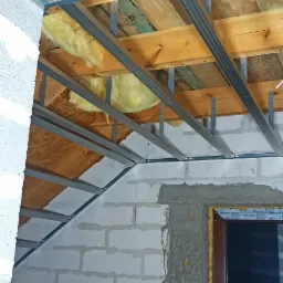 Poddasze w trakcie ocieplania: widoczne drewniane elementy konstrukcyjne, metalowy stelaż, wełna izolacyjna, ściany z białych bloczków, okno osadzone w surowym otworze.