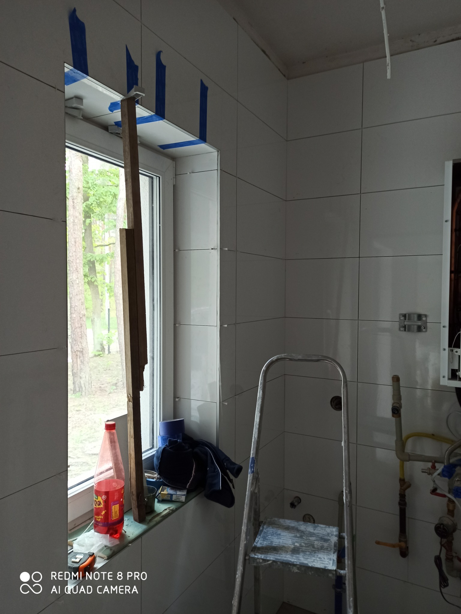 Ściana łazienki w trakcie wykańczania, z białymi płytkami, oknem zabezpieczonym taśmą malarską, opartą deską, butelką napoju, ubraniem na parapecie i aluminiową drabiną.