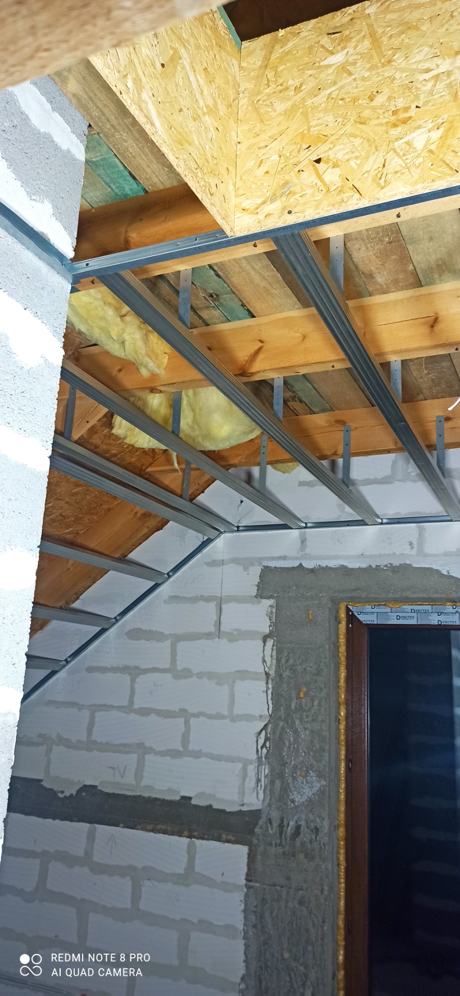 Poddasze w trakcie ocieplania: widoczne drewniane elementy konstrukcyjne, metalowy stelaż, wełna izolacyjna, ściany z białych bloczków, okno osadzone w surowym otworze.