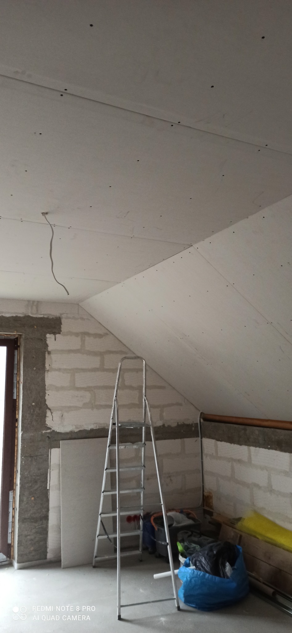 Poddasze w trakcie prac wykończeniowych: widoczne płyty kartonowo-gipsowe na skosach, aluminiowa drabina, fragment ściany z bloczków oraz wystający przewód elektryczny.