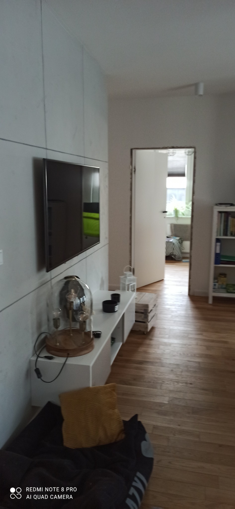 Salon z drewnianą podłogą, szarą ścianą z zamontowanym telewizorem, białą szafką RTV i otwartymi drzwiami do innego pomieszczenia.