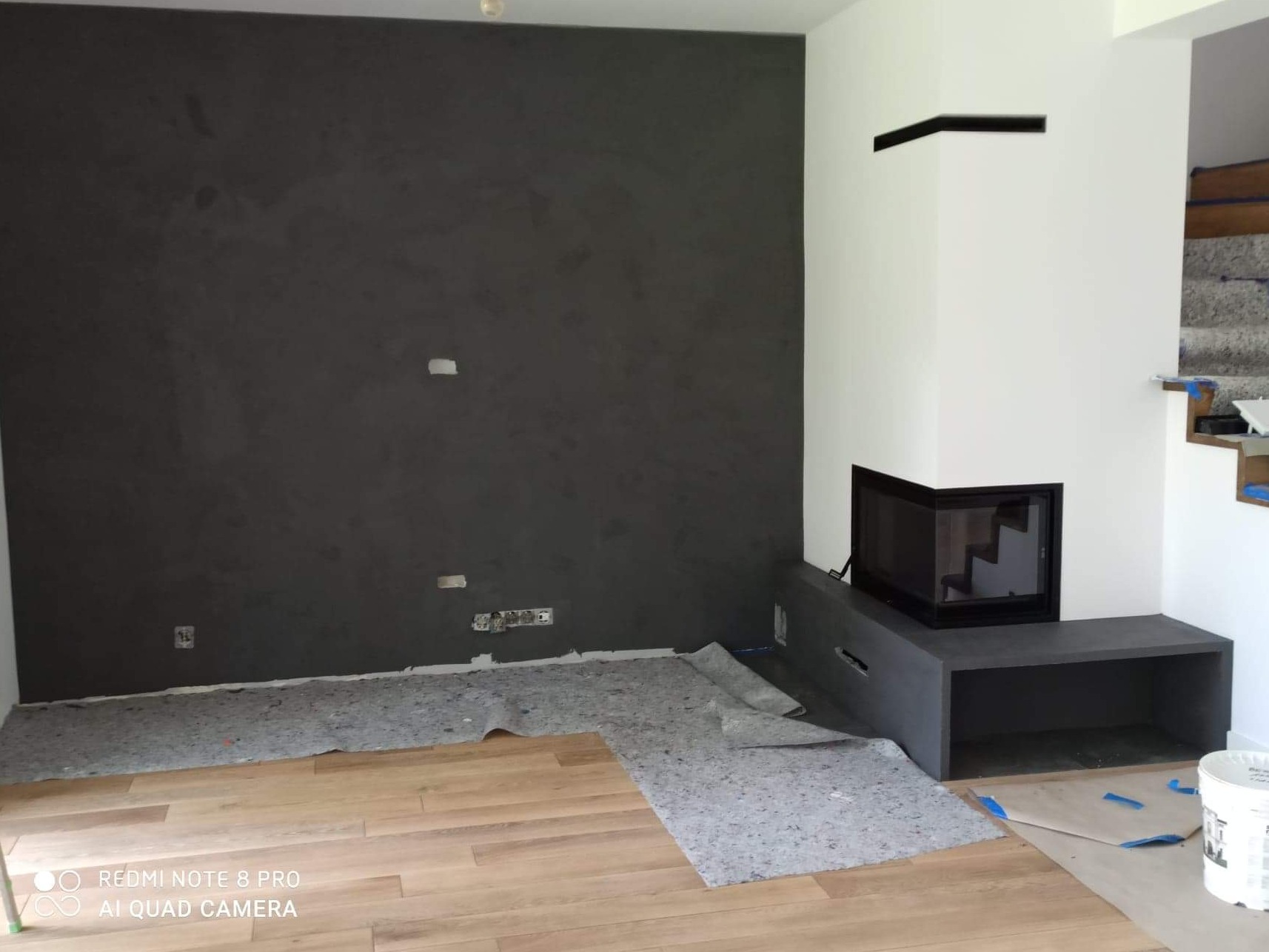 Pokój w trakcie prac wykończeniowych: ściana pokryta ciemnoszarym tynkiem dekoracyjnym, częściowo zabezpieczona podłoga panelowa, biały kominek z czarnym wkładem kominkowym.