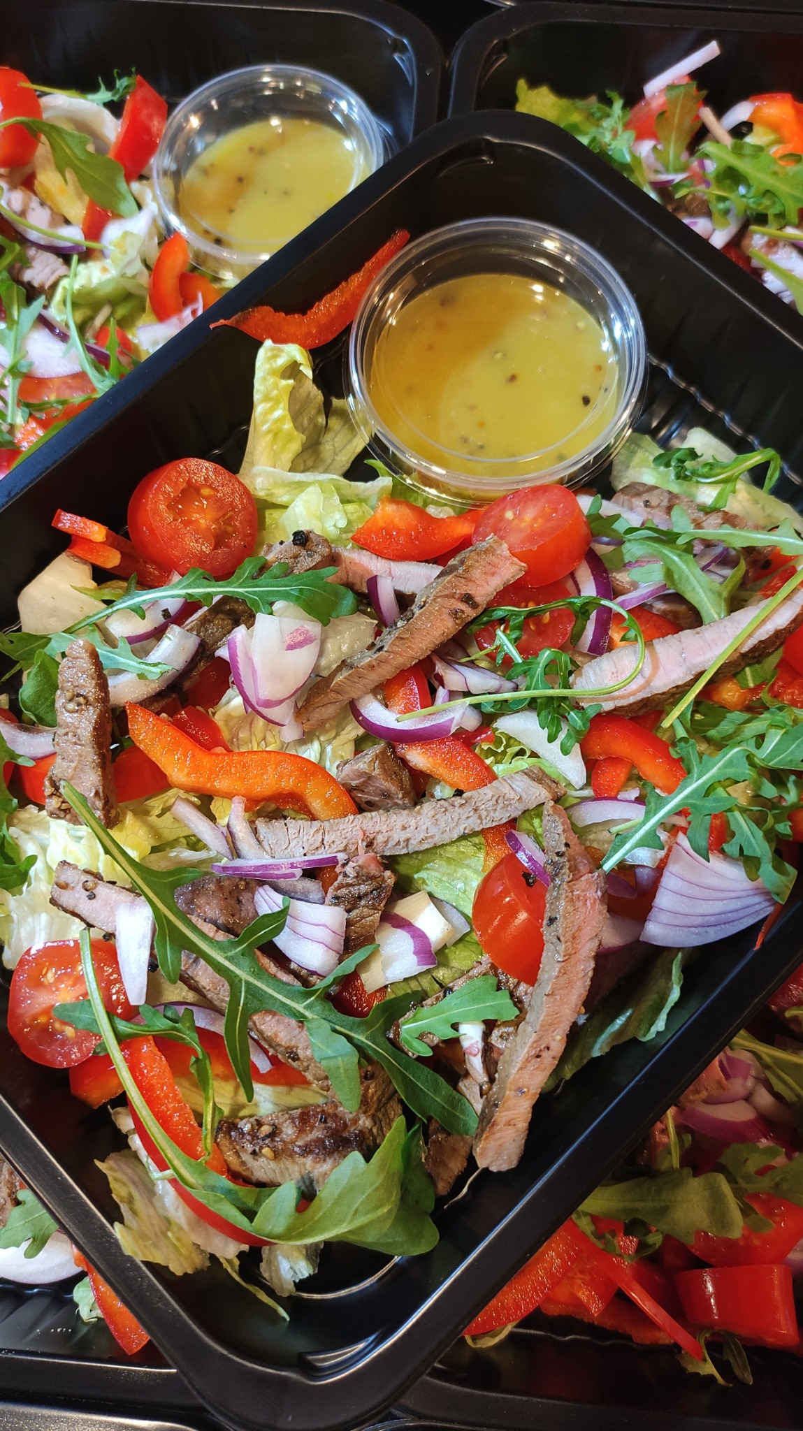 Sałatka z grillowaną wołowiną, pomidorami, czerwoną cebulą, papryką i rukolą w czarnym pojemniku z sosem w osobnym kubeczku.