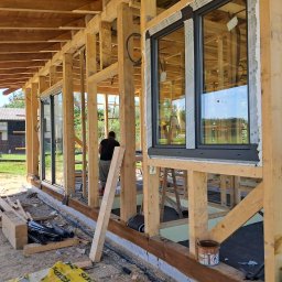 MARCIN KOZAK MARKER - Montaż okien PCV w surowym drewnianym domu. Widoczne ramy okienne Salamander i elementy konstrukcyjne. Etap budowy, okna częściowo zabezpieczone folią.