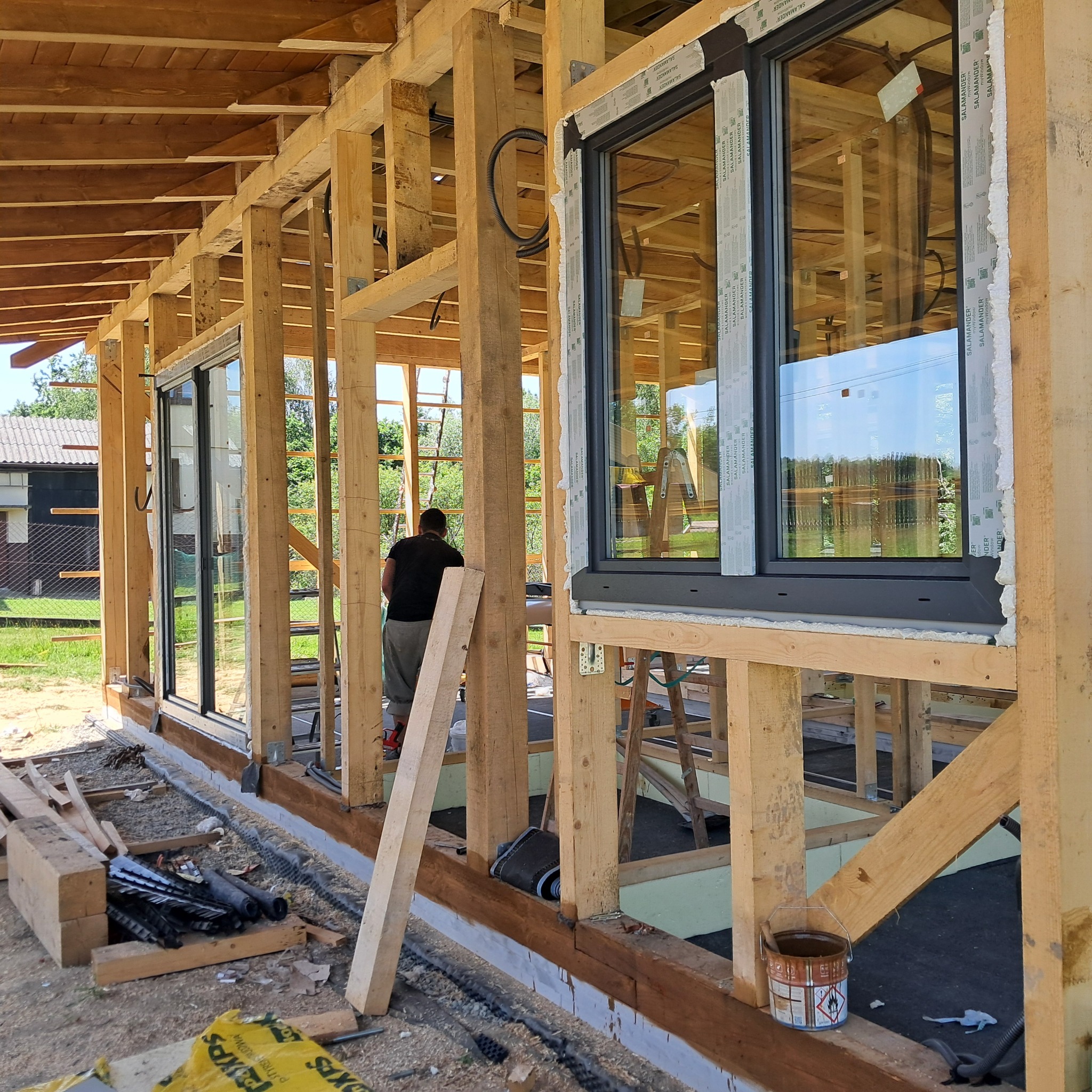 Montaż okien PCV w surowym drewnianym domu. Widoczne ramy okienne Salamander i elementy konstrukcyjne. Etap budowy, okna częściowo zabezpieczone folią.