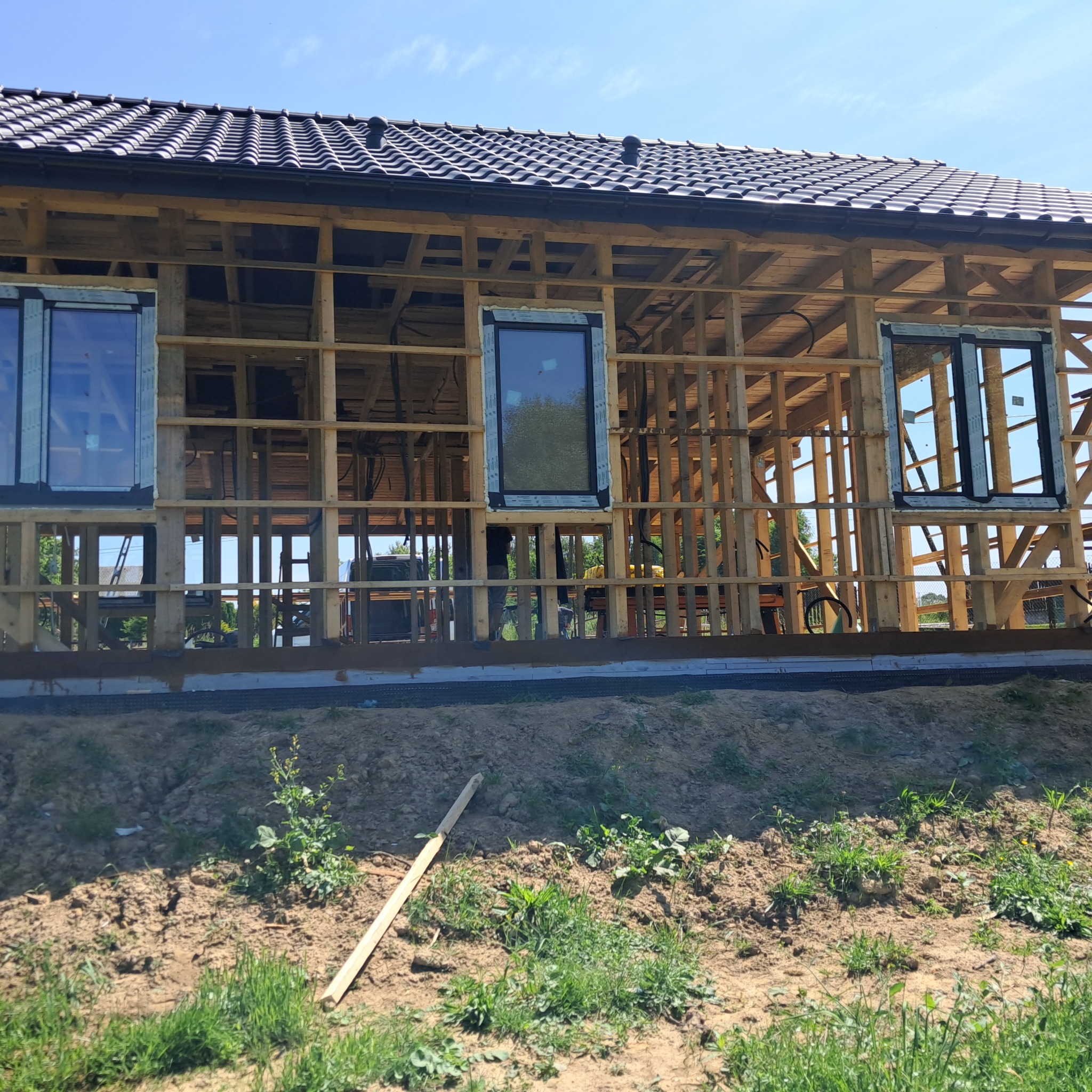 Montaż okien w surowym budynku z drewnianą konstrukcją szkieletową i czarnym dachem. Widoczne trzy okna z ramami. Na pierwszym planie ziemia z roślinnością.