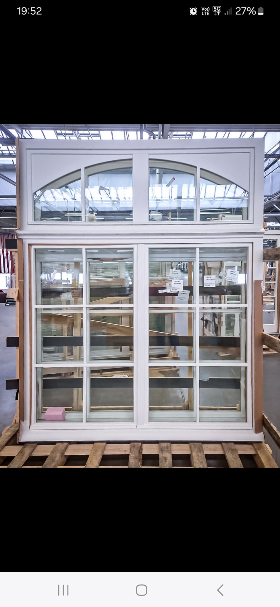 Białe okno PCV z górnym łukowym panelem, ustawione na palecie w magazynie, widoczne etykiety i inne okna w tle. Nowoczesny design, jasne wnętrze.