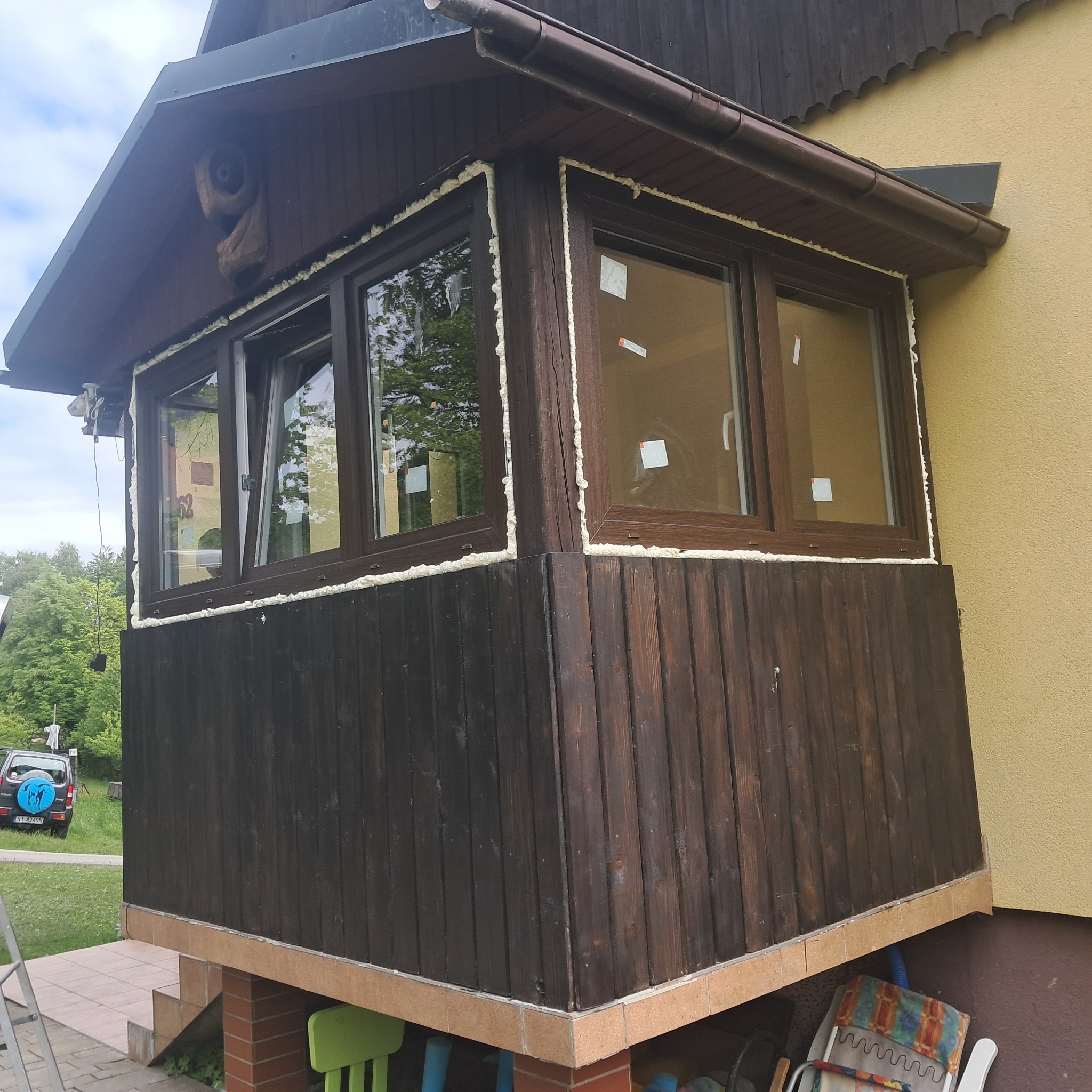 Zabudowany balkon z brązowymi oknami, uszczelnionymi pianką montażową. Widoczna drewniana konstrukcja i fragment elewacji budynku w odcieniu żółci.