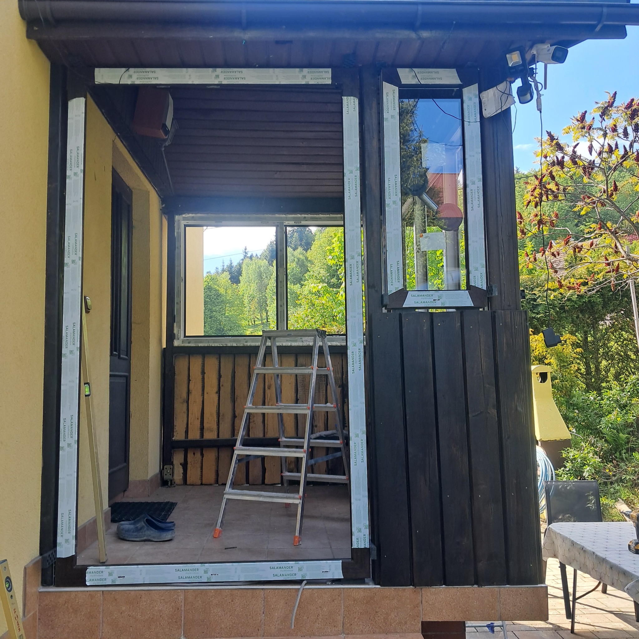 Montaż okien PCV marki Salamander w drewnianej zabudowie tarasu. Widoczna drabina, poziomica i narzędzia. W tle zieleń drzew, błękit nieba. Etap prac.