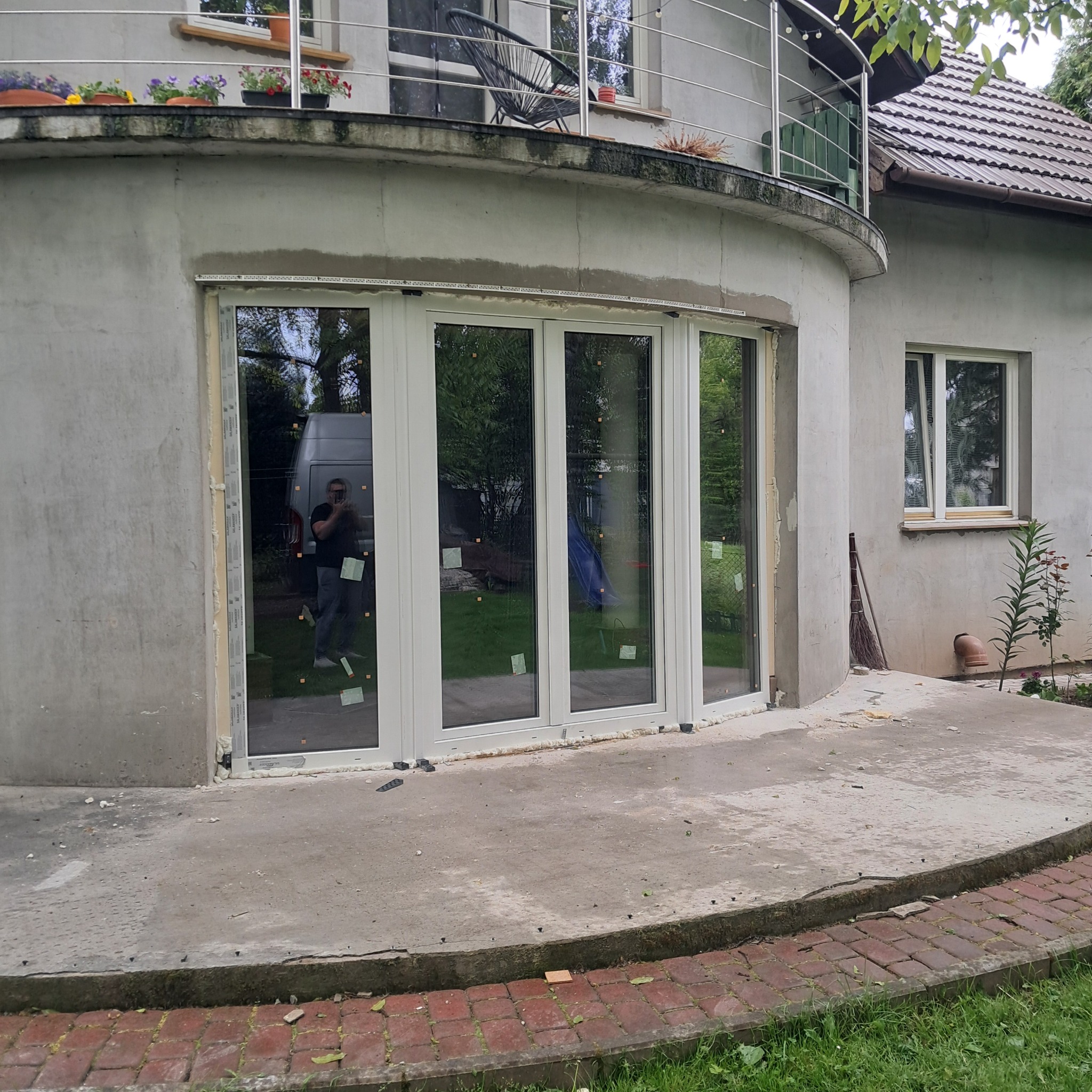 Montaż białych okien PCV w zaokrąglonej ścianie domu. Widoczne pianowanie i odbicie osoby robiącej zdjęcie. Na górze balkon z kwiatami.