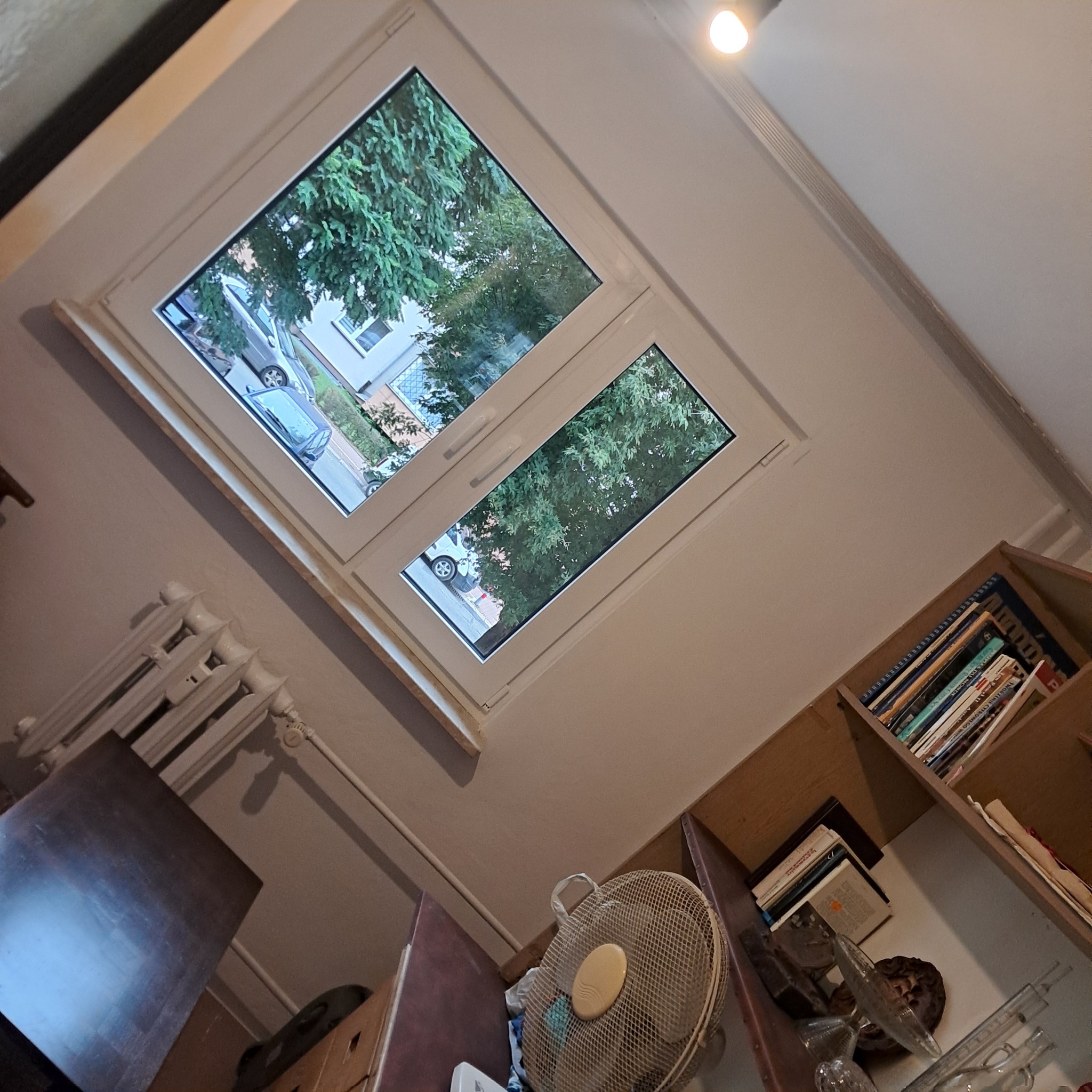 Białe okno PCV z widokiem na zieleń i ulicę, widziane z perspektywy wnętrza pokoju z grzejnikiem i regałem. Nietypowy kąt ujęcia.