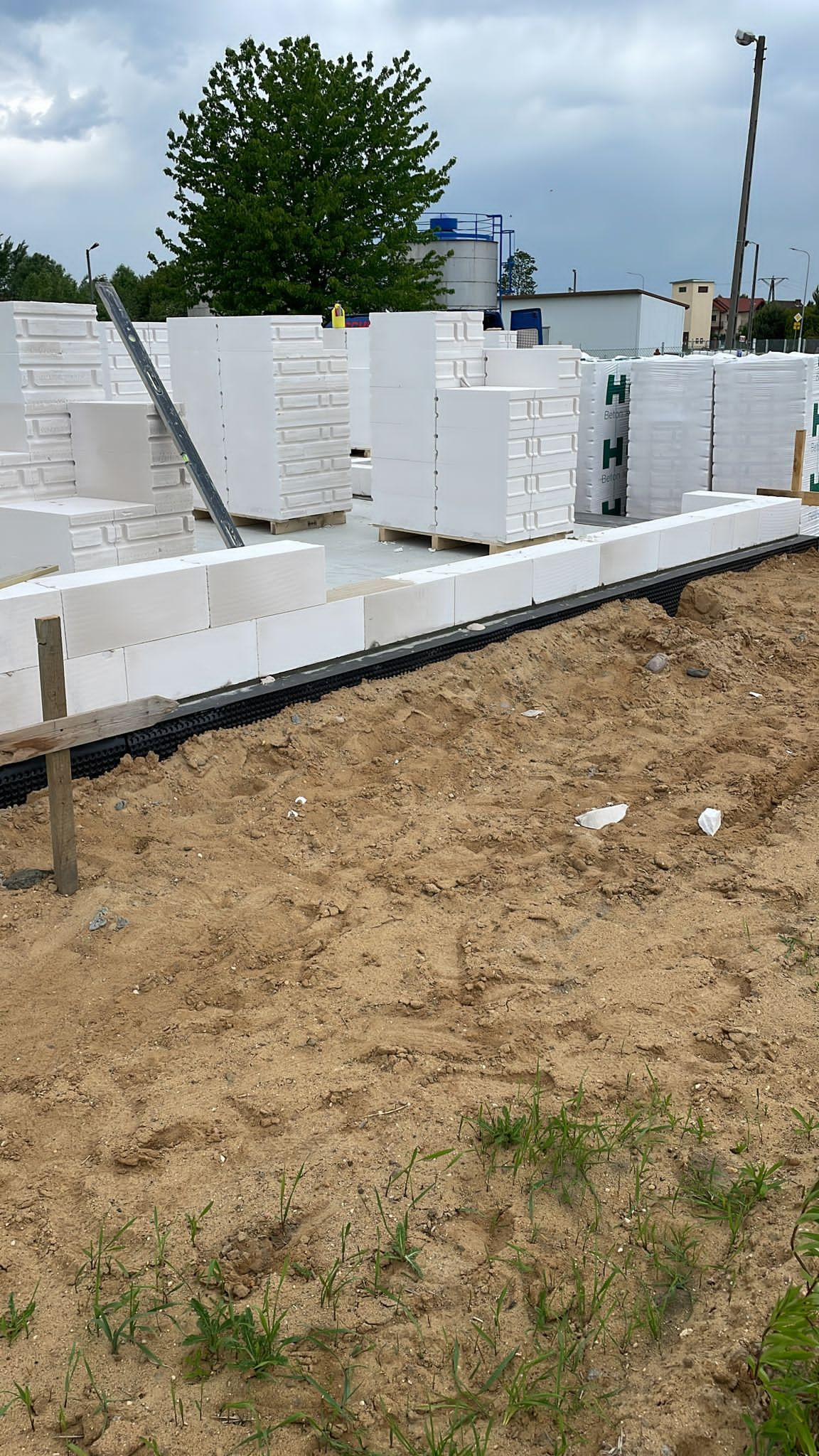 Wznoszona ściana z białych bloczków na fundamencie; widoczna izolacja pozioma i palety z materiałem budowlanym w tle.