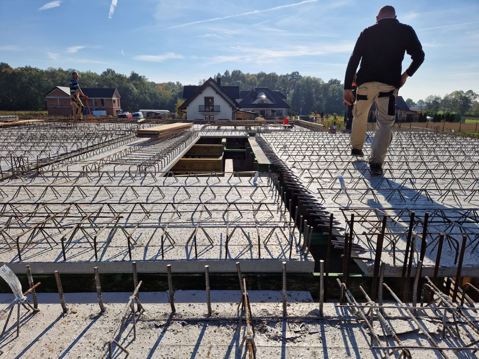 Zbrojenie płyty fundamentowej z widocznymi elementami dystansowymi i wystającymi prętami, dwóch pracowników w oddali, domy w tle.