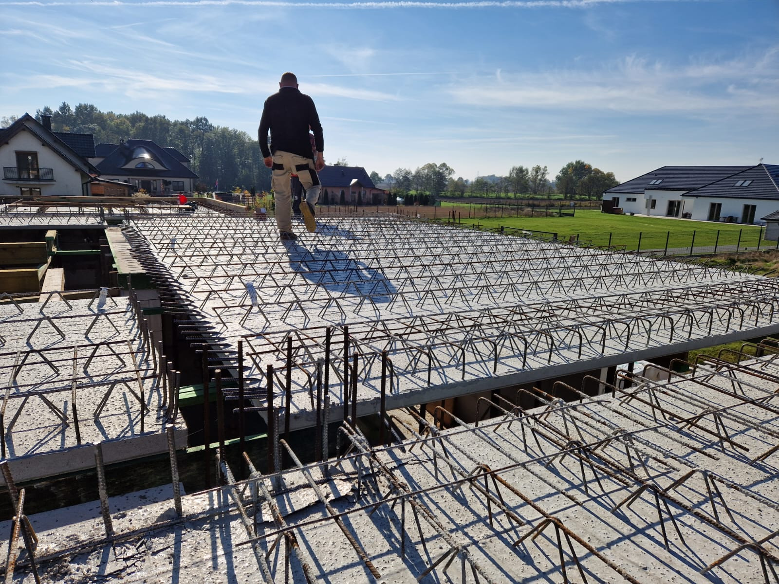Prace zbrojarskie na budowie domu jednorodzinnego, widok z góry na siatkę ze stali zbrojeniowej, w tle budynki mieszkalne i błękitne niebo.