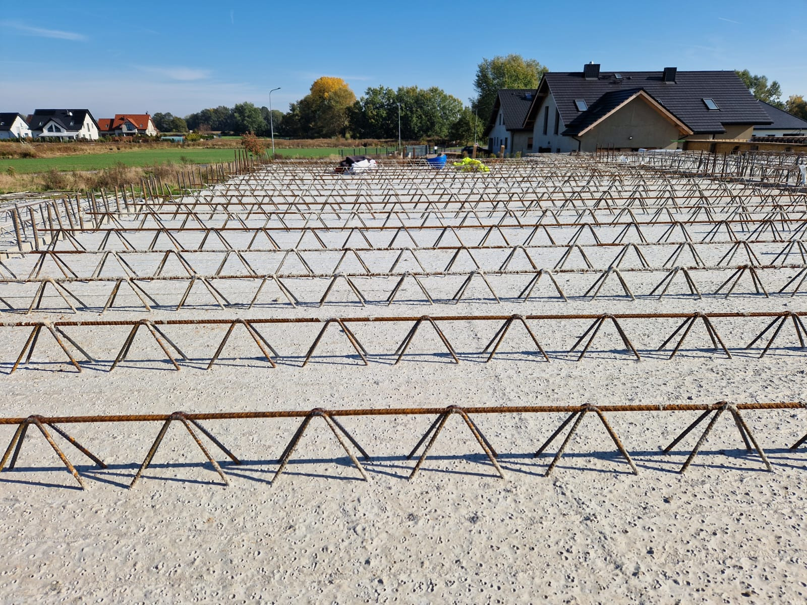 Zbrojenie płyty fundamentowej z widocznymi stalowymi prętami ułożonymi w trójkątny wzór, na tle częściowo ukończonego budynku mieszkalnego i błękitnego nieba.