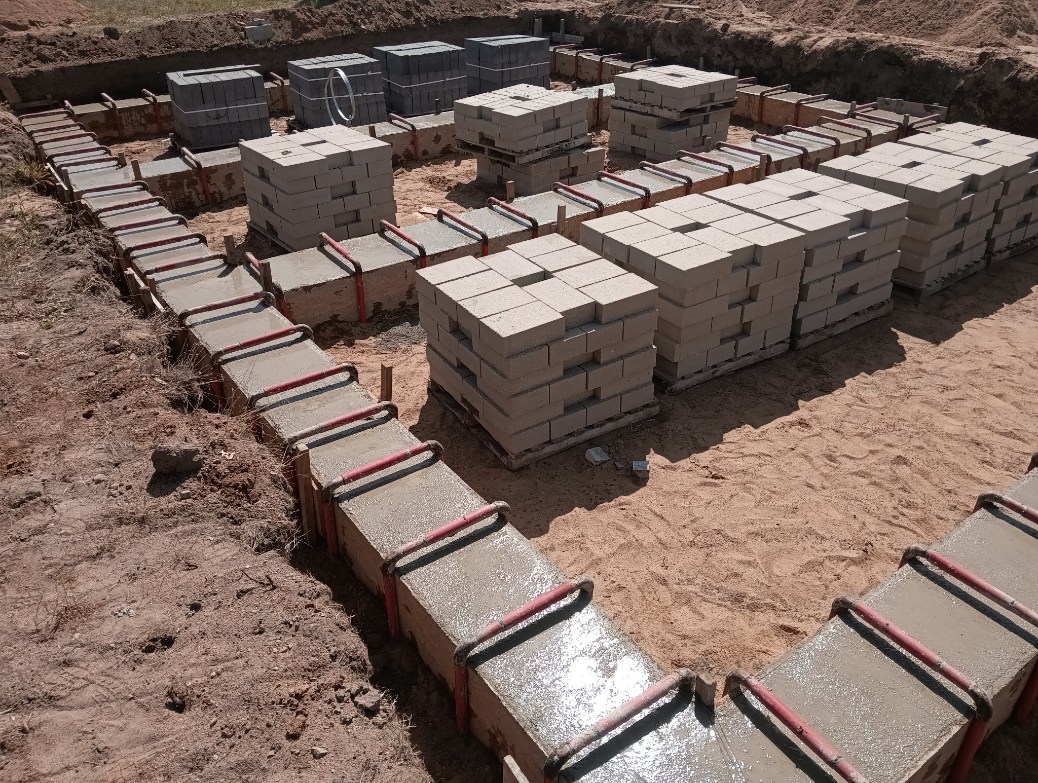 Zalane betonem szalunki fundamentowe z czerwonymi wspornikami, obok ułożone bloczki betonowe. Widok z góry na wykopany teren budowy.