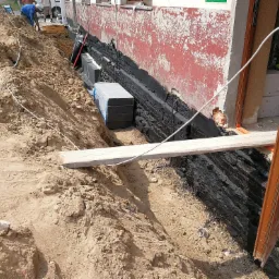 Prace ziemne przy fundamencie budynku z widoczną warstwą hydroizolacji, zabezpieczoną styropianem i folią, oraz wykopem wypełnionym ziemią.