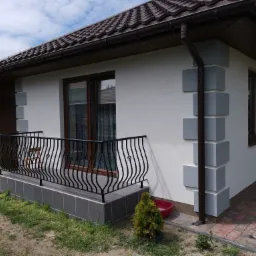 Jasny dom jednorodzinny z brązowym dachem i balkonem z czarną, kutą balustradą, udekorowany szarymi elementami architektonicznymi na elewacji.