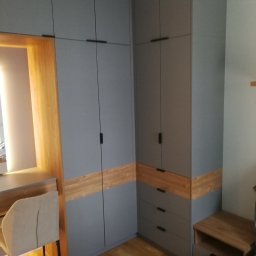 Mirosław M. Mróz mgr sztuki projektant wnętrz stolarz - Szara szafa na wymiar z drewnianym akcentem, dopasowana do biurka i szafki nocnej, tworząca spójny design wnętrza. Minimalistyczne uchwyty i drewniana podłoga.