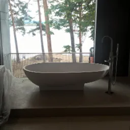 Nowoczesna, wolnostojąca wanna w surowym, minimalistycznym wnętrzu łazienki z widokiem na jezioro przez duże okno. Elementy w trakcie wykończenia, zabezpieczone folią.