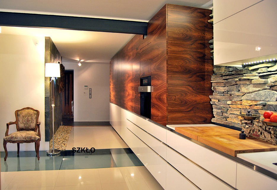 Nowoczesna kuchnia z frontami z ciemnego drewna o wyraźnym usłojeniu, białymi szafkami, kamienną ścianą i drewnianym blatem, oświetlona listwą LED pod szafkami wiszącymi. W tle widoczny fotel...