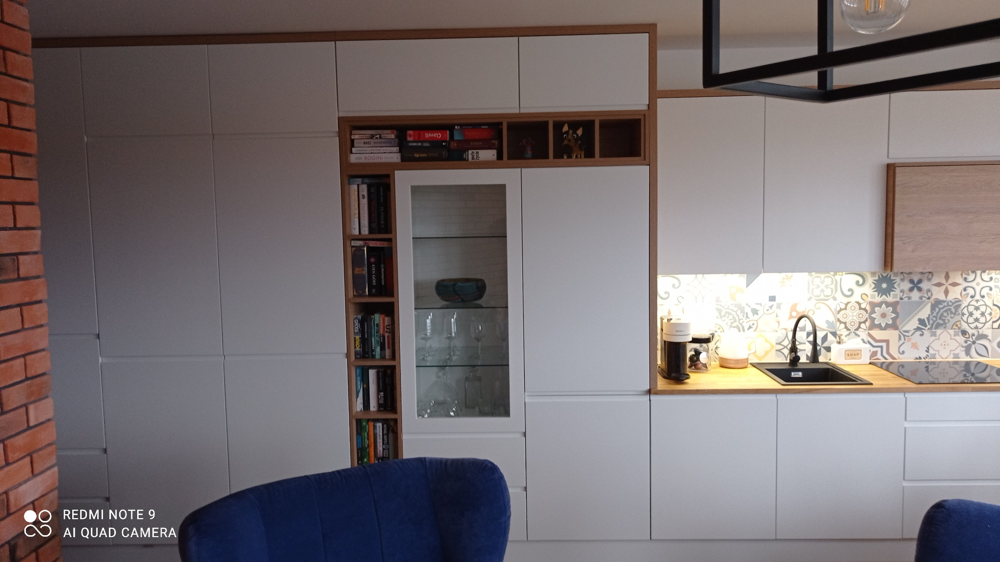 Biała zabudowa kuchenna na wymiar z drewnianymi akcentami, widoczna szklana witryna z książkami i dekoracjami, wzorzyste płytki nad blatem.