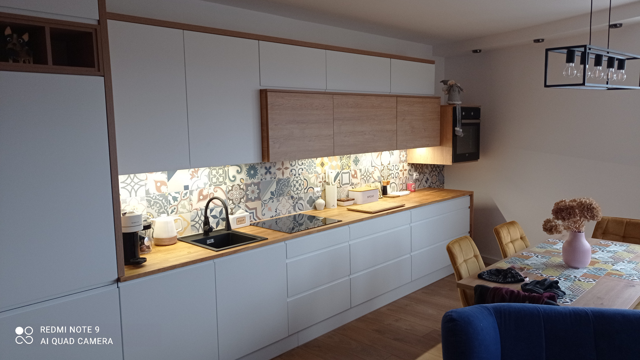 Nowoczesna kuchnia z białymi szafkami, drewnianym blatem i wzorzystą mozaiką nad blatem. Widoczny czarny zlew i płyta grzewcza. Oświetlenie LED pod szafkami.