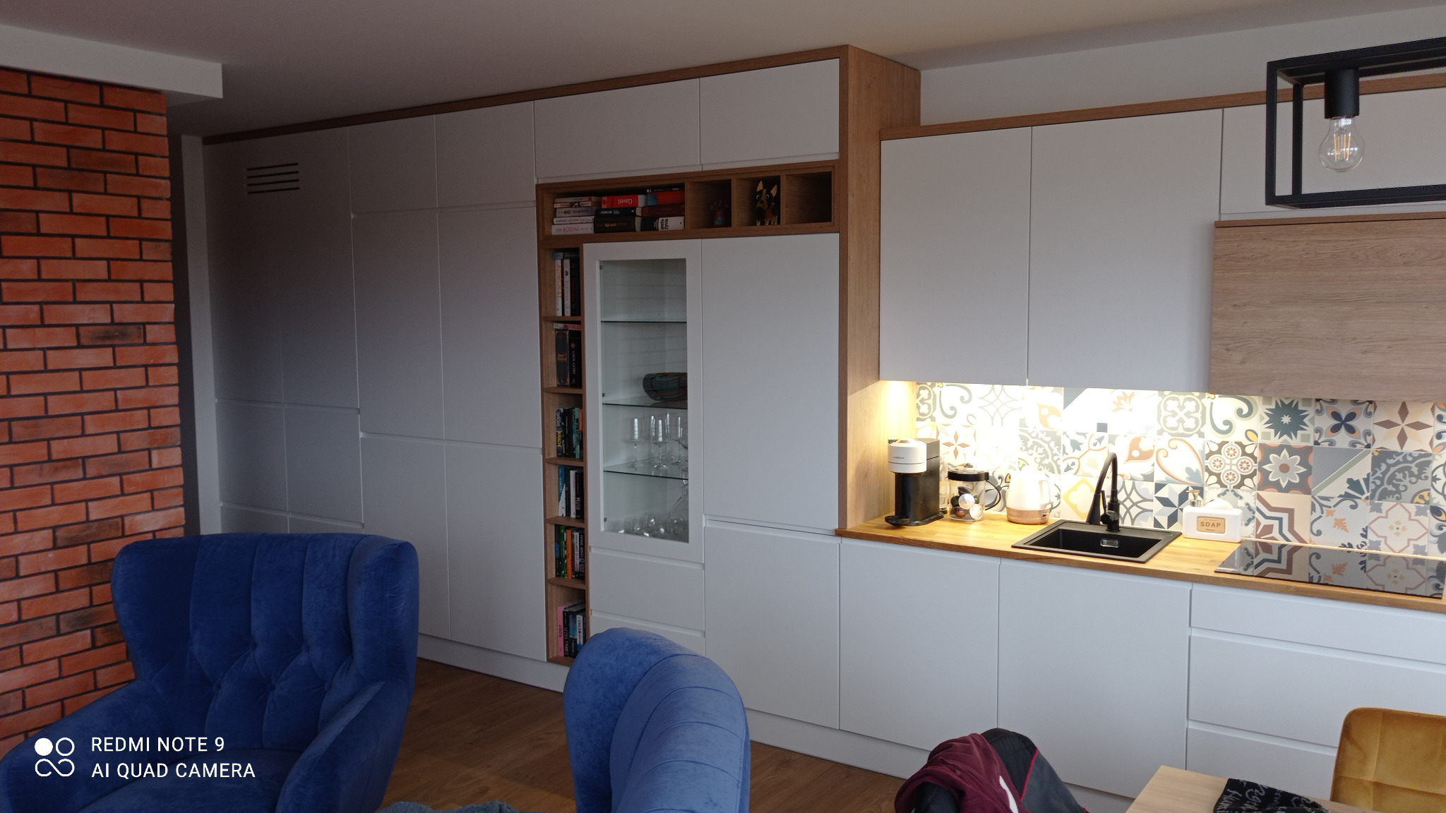 Biała kuchnia na wymiar z drewnianymi akcentami i wzorzystą ścianą nad blatem. W tle ceglana ściana i niebieskie fotele, tworzące kontrastowe, nowoczesne wnętrze.