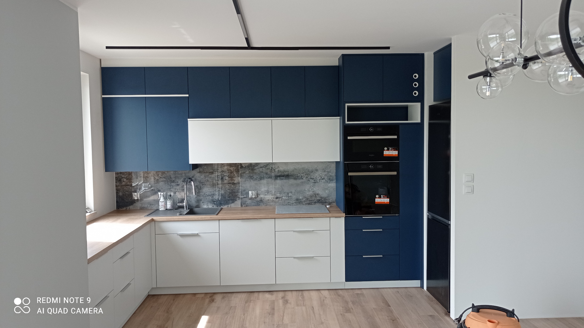 Nowoczesna kuchnia z białymi dolnymi szafkami i granatowymi górnymi, drewniany blat, szara ściana nad blatem, oświetlenie sufitowe i sprzęt AGD w zabudowie.