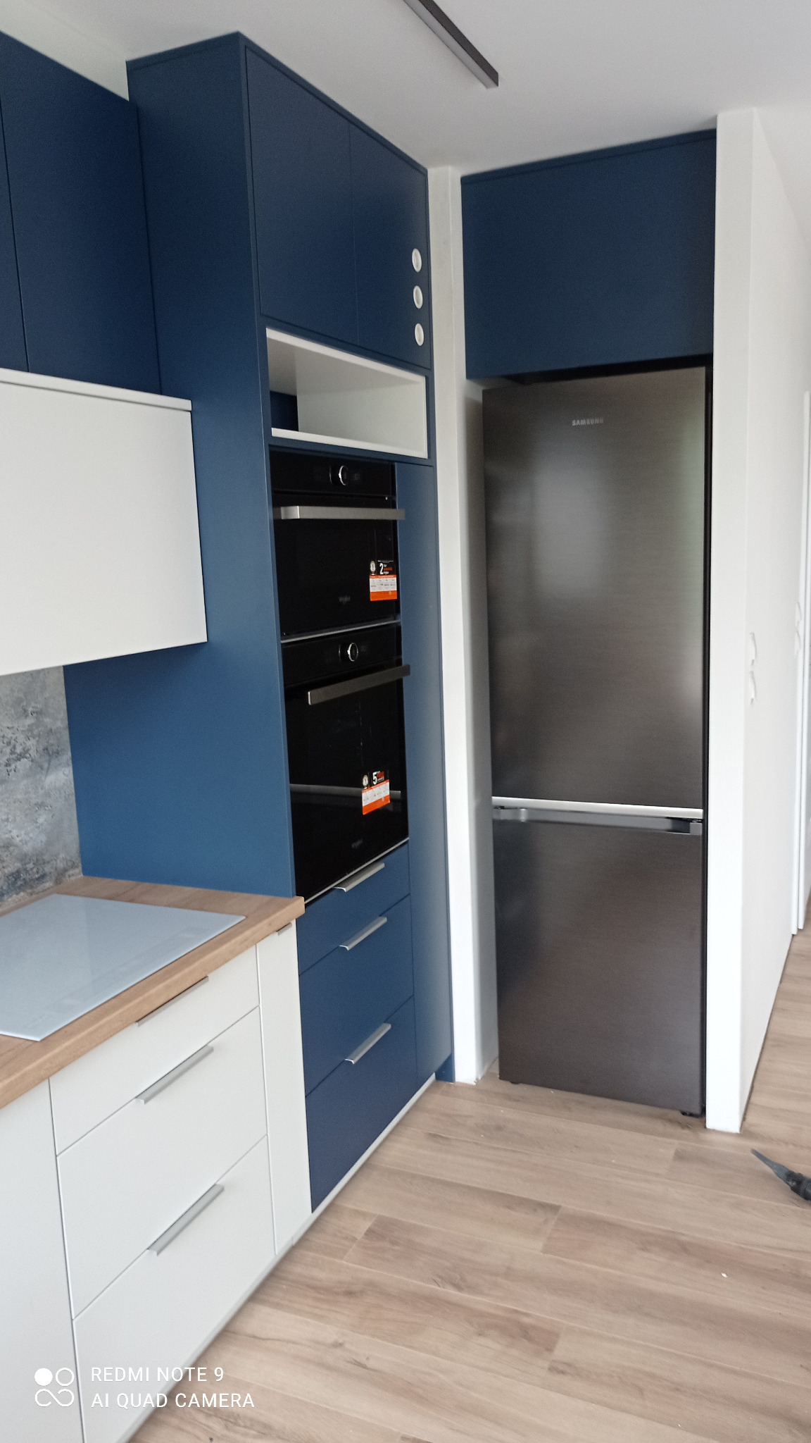 Nowoczesna kuchnia w odcieniach bieli i granatu. Zabudowa kuchenna z piekarnikami, szufladami i szafkami. Na blacie płyta indukcyjna. Obok zabudowana lodówka.