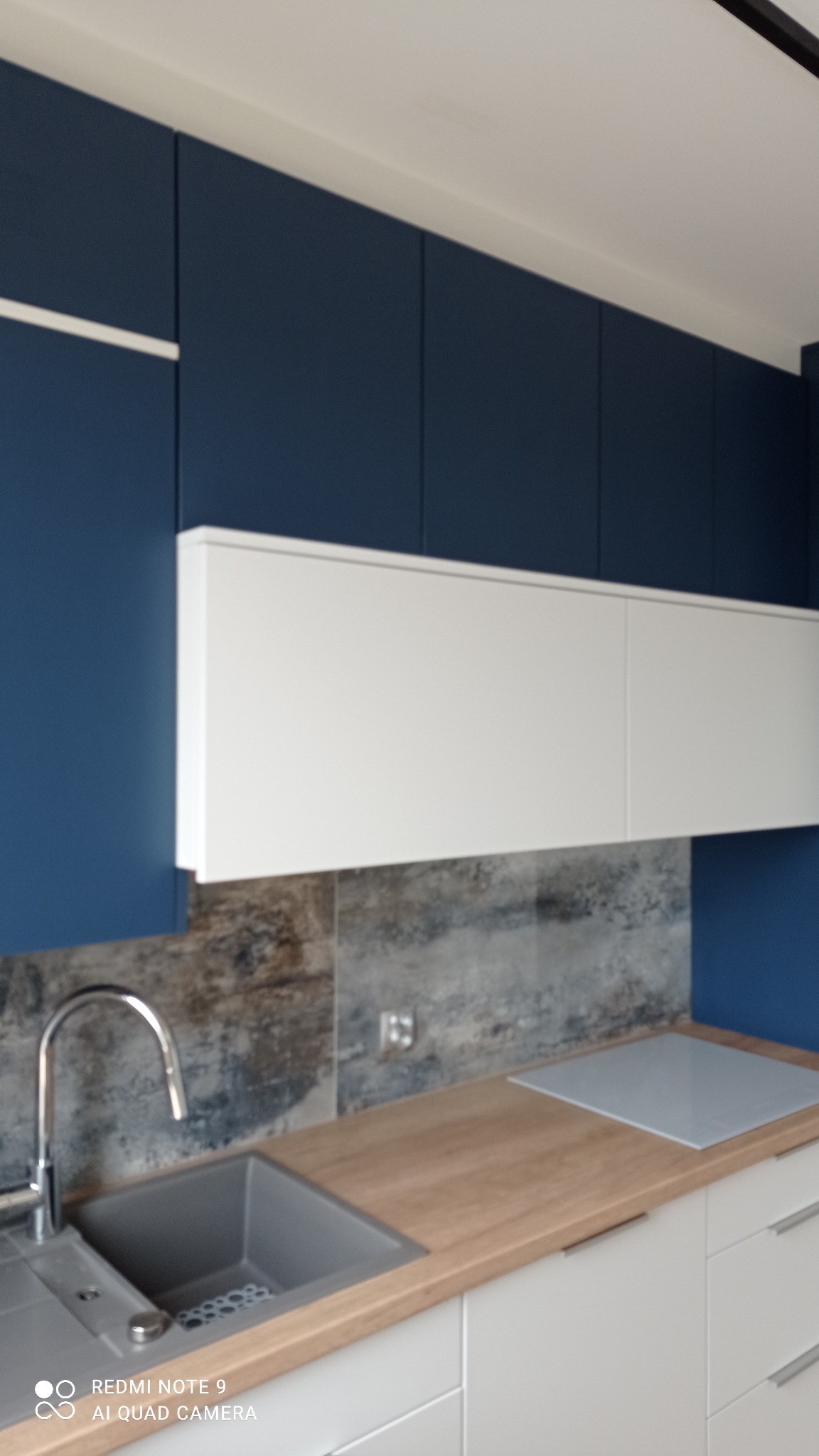 Nowoczesna kuchnia z granatowymi szafkami górnymi i białymi dolnymi. Blat drewnopodobny, szary zlew i bateria. Płytki imitujące kamień na ścianie.